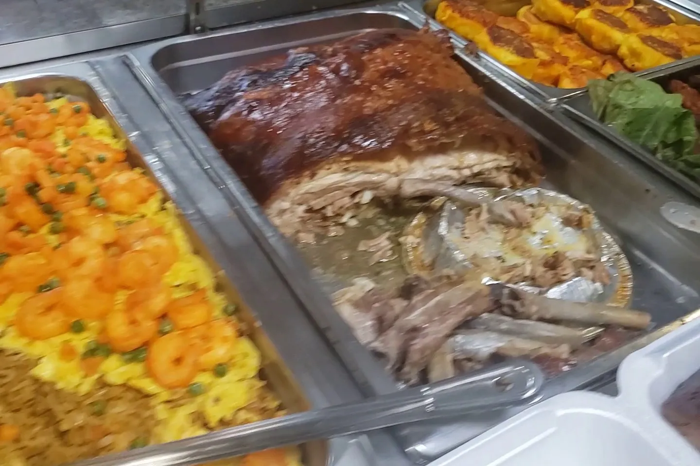 Steam table trays of shrimp with peas over yellow rice, a whole roast pork leg, and golden griddled patties